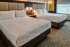 A hotel room with two beds, a headboard with built-in lighting, a nightstand with a lamp, and a large window with a view of a parking lot.