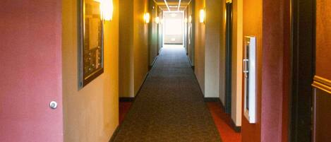 A hotel hallway with carpeted floors, doors on both sides, and wall-mounted light fixtures.