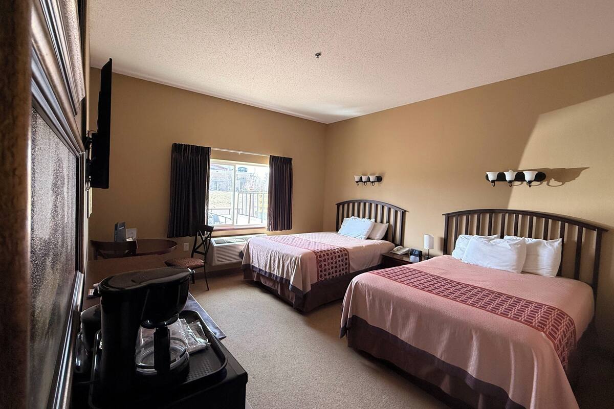 A hotel room with two beds, a desk with a coffee maker, a chair, a window with curtains, and a wall-mounted light fixture.
