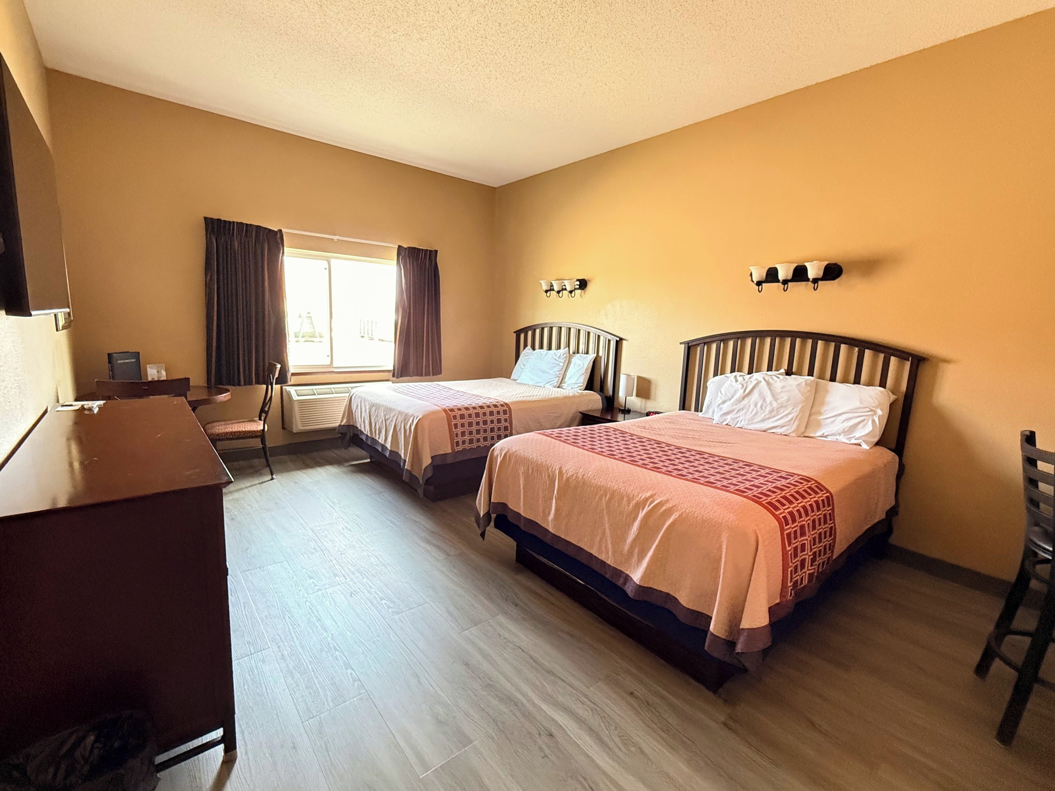 A hotel room with two beds, a desk, a chair, a window with curtains, and wall-mounted lights.
