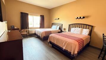 A hotel room with two beds, a desk, a chair, a window with curtains, and wall-mounted lights.