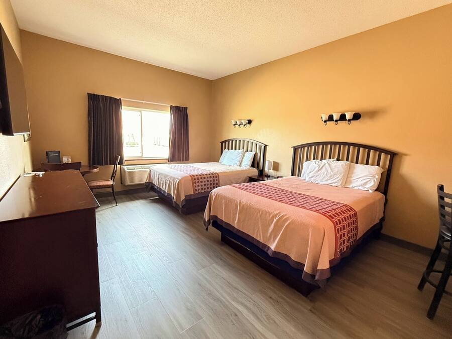 A hotel room with two beds, a desk, a chair, a window with curtains, and wall-mounted lights.