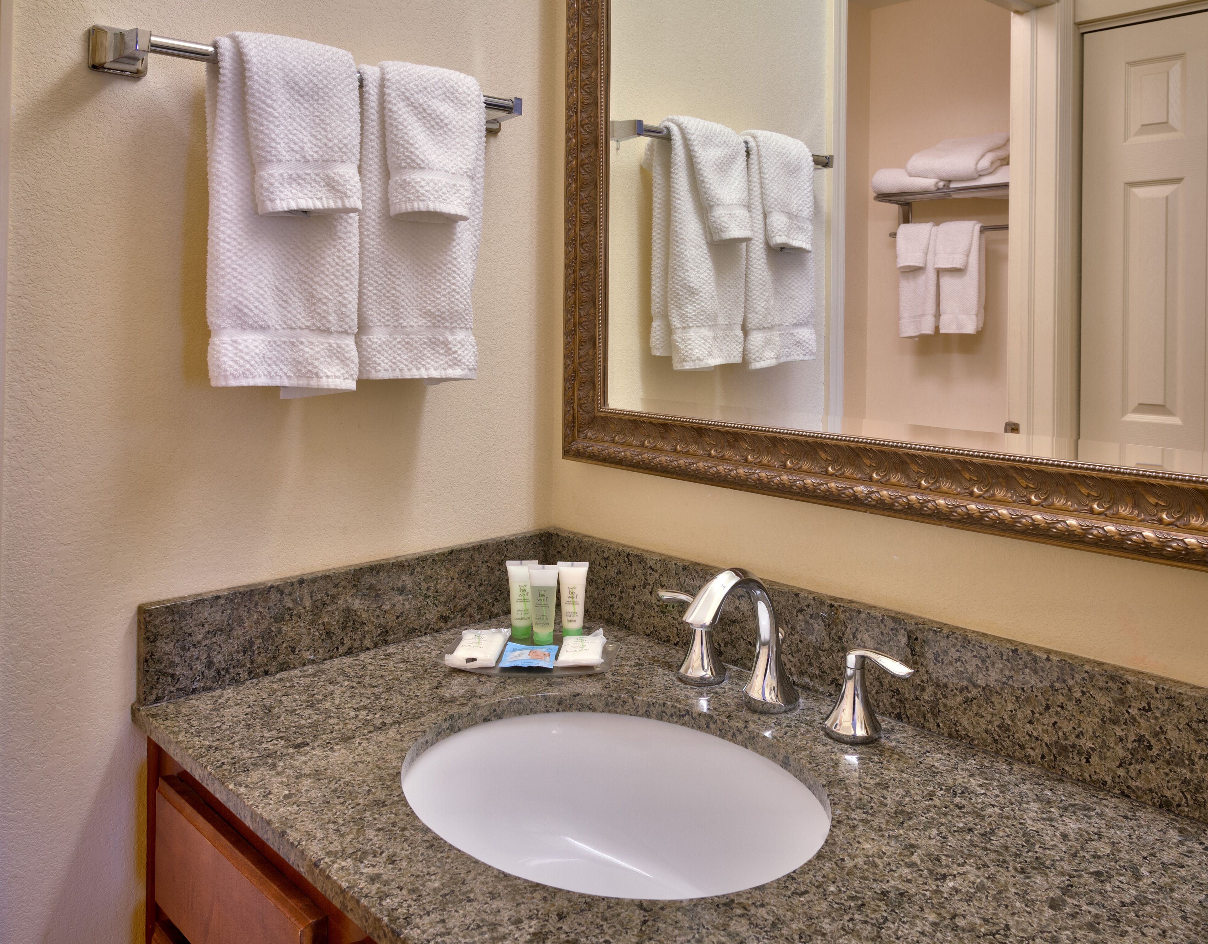 A bathroom with a granite countertop, a white sink, two faucets, and a mirror with a gold frame.
