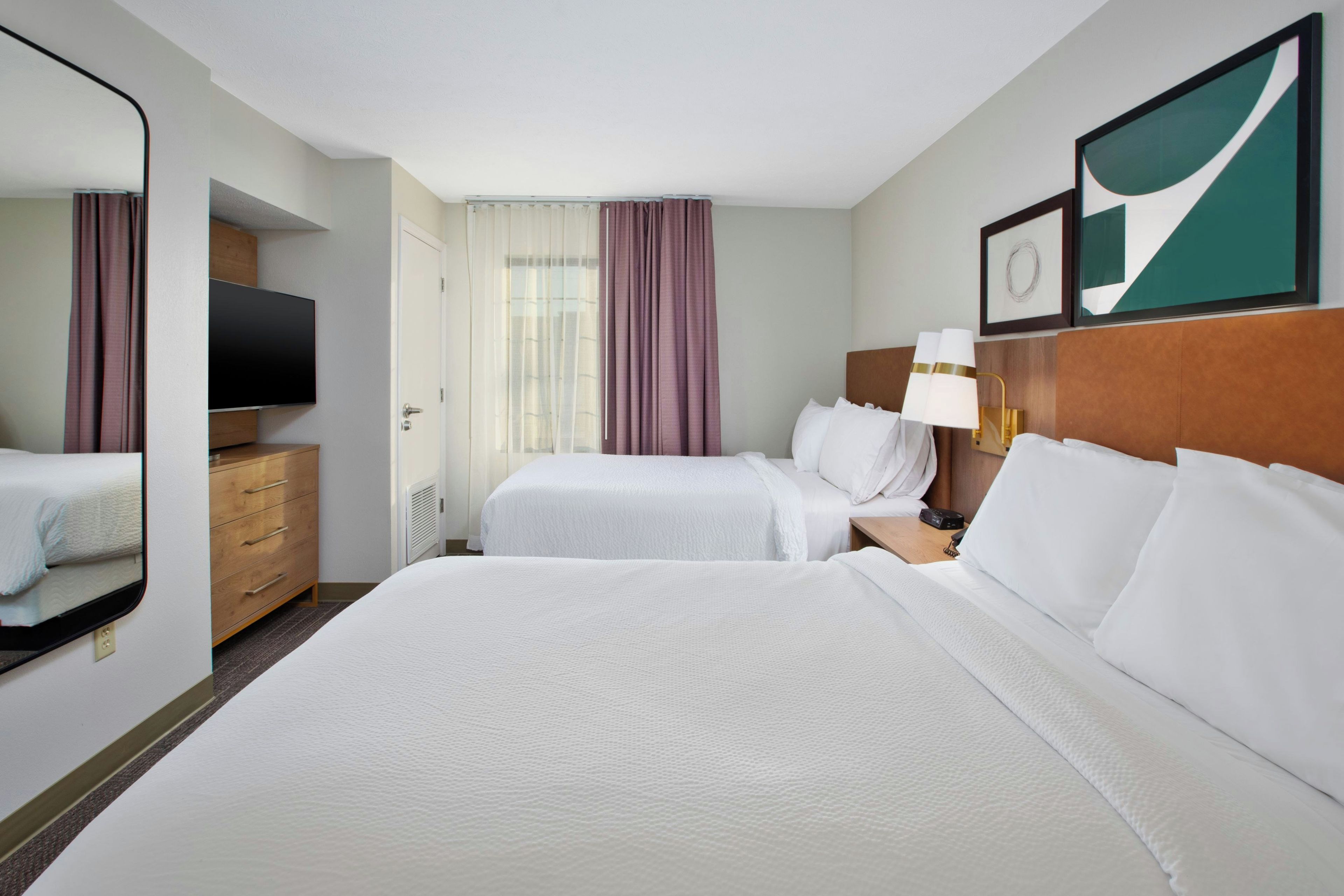 A hotel room with two beds, a TV, a dresser, and a window with curtains.
