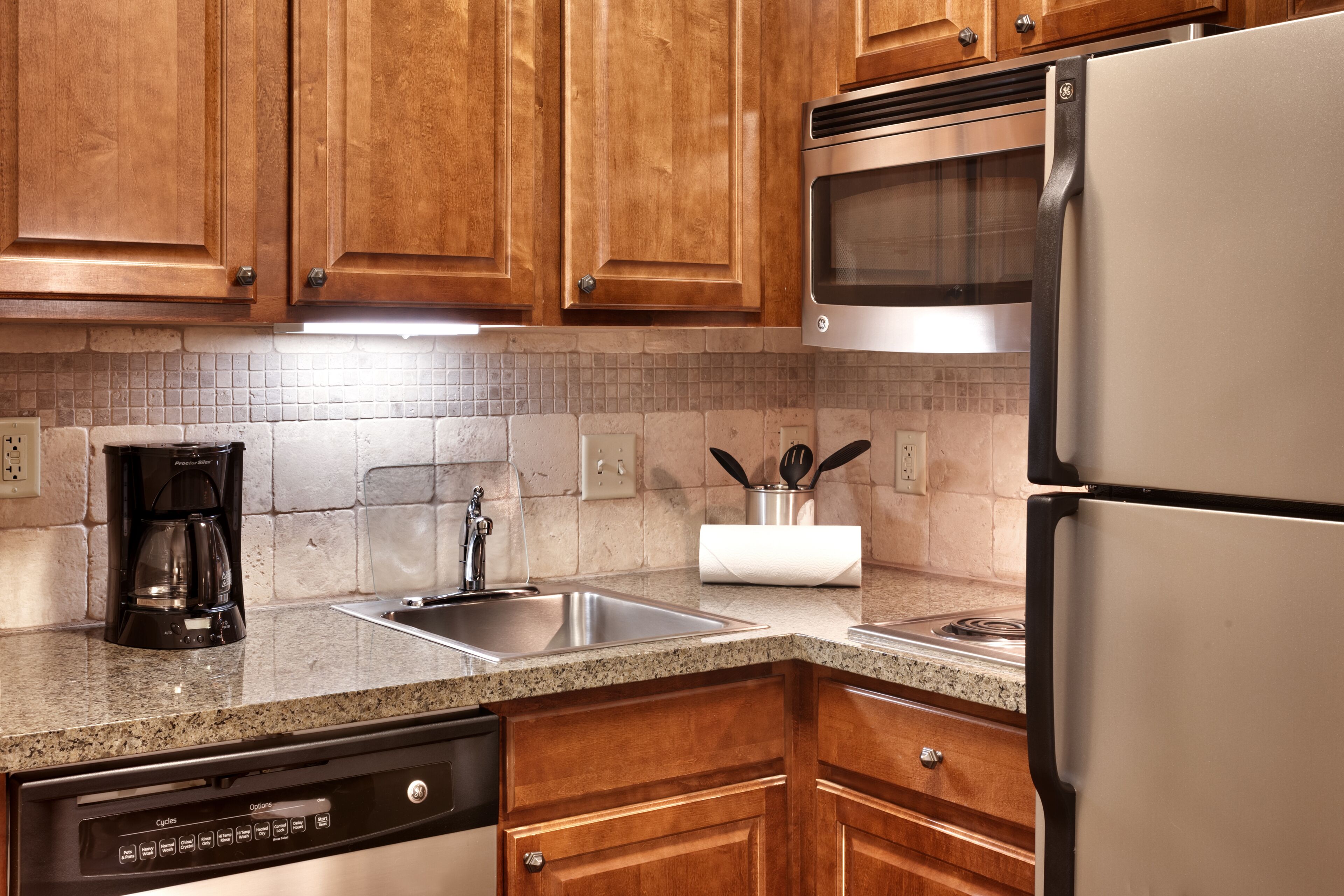 Una cocina con gabinetes de madera, un refrigerador de acero inoxidable, un microondas integrado, una cafetera y un lavamanos.