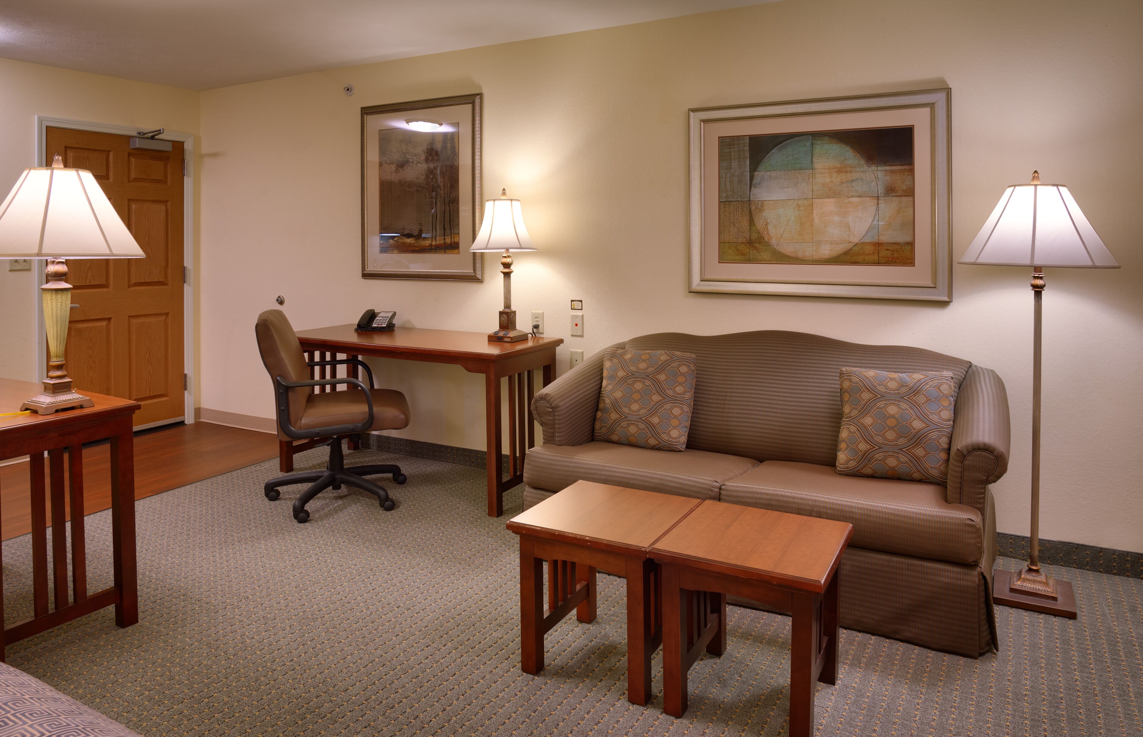 A hotel room with a sofa, a desk, a chair, two lamps, and two framed artworks on the walls.