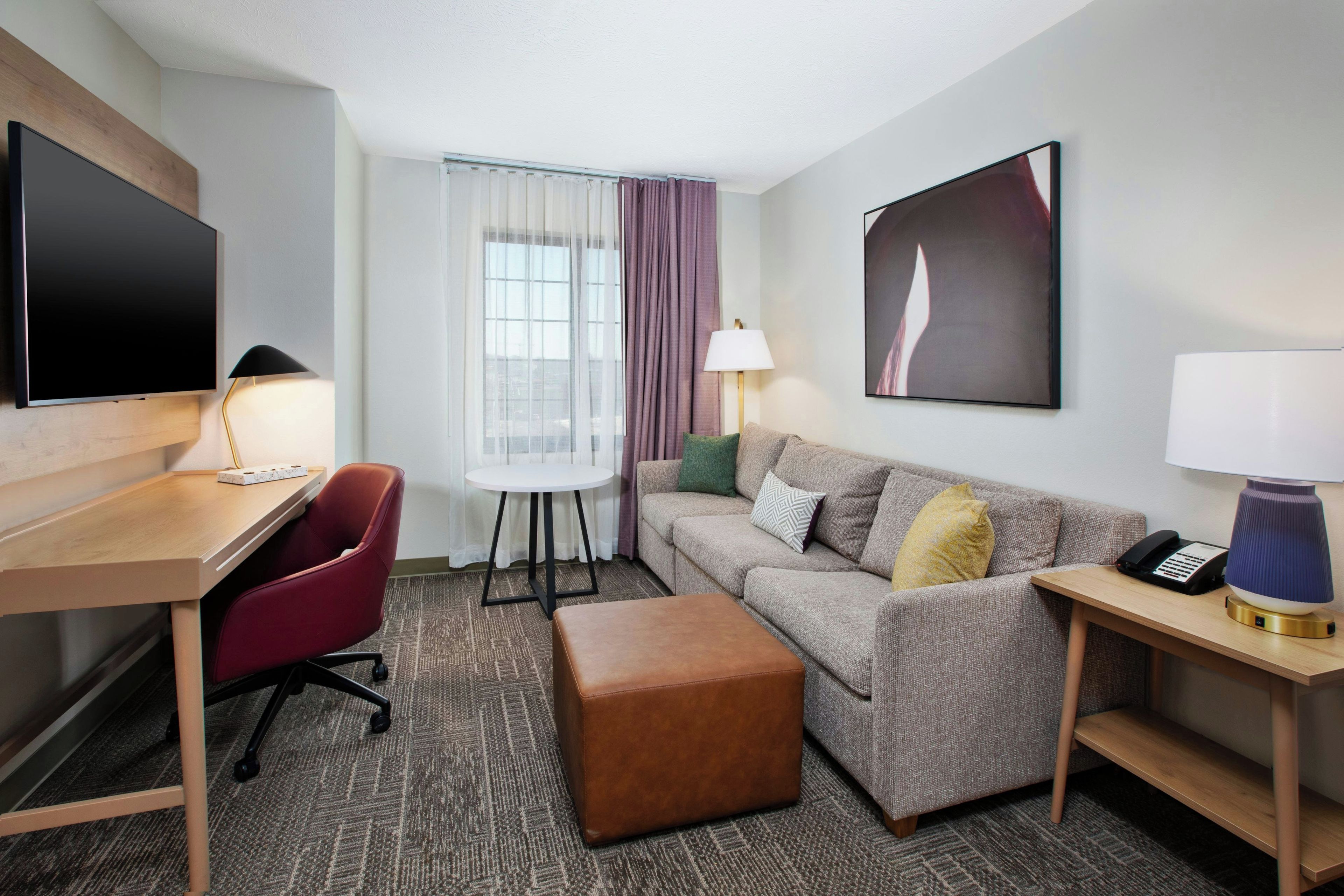 Habitación de hotel con televisión de pantalla plana, escritorio, una silla de oficina roja, sofá con cojines, un reposapiés pequeño, lámpara, teléfono y ventana con cortinas.
