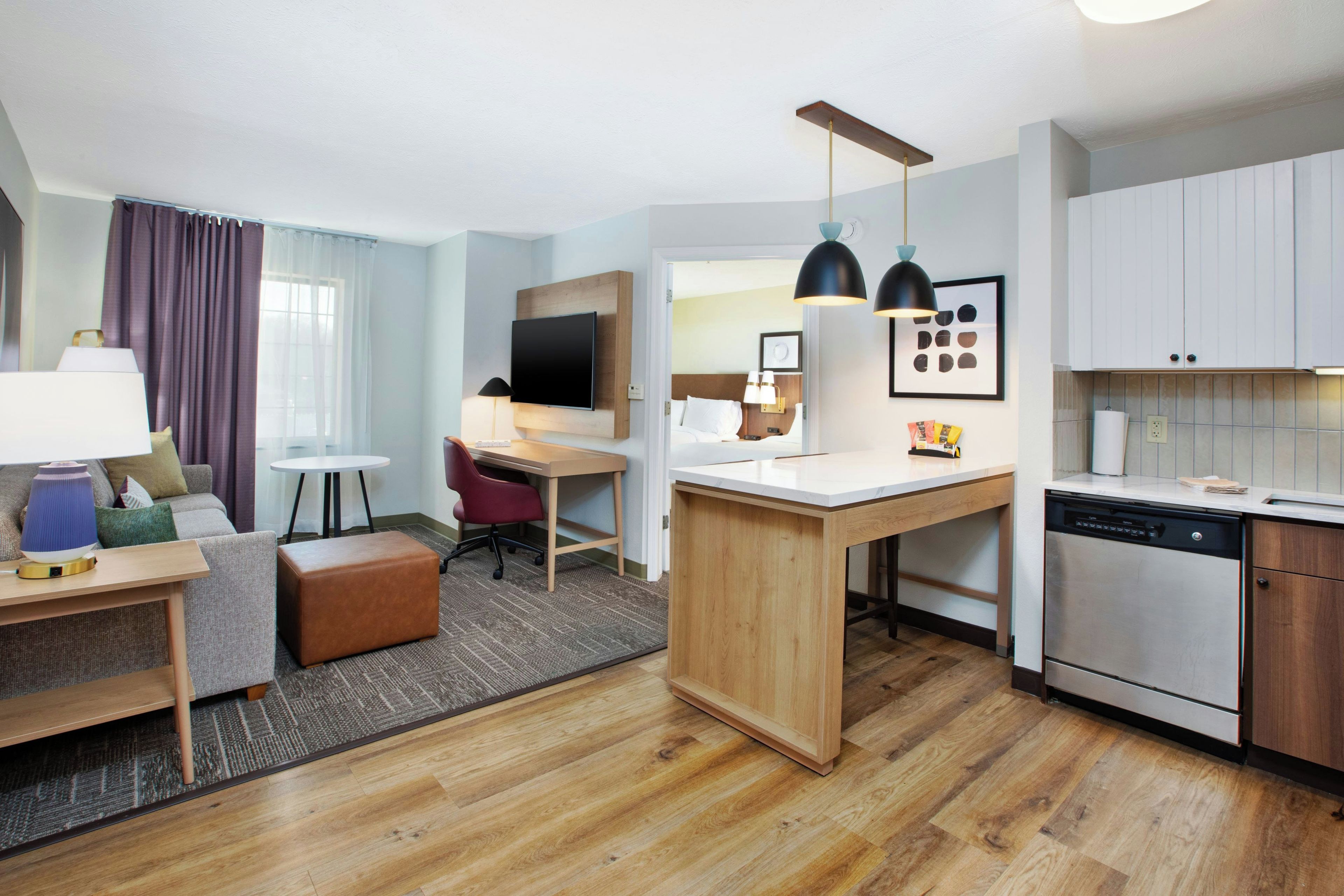 A modern hotel room with a wooden floor, a kitchenette, a dining area, and a living space with a sofa and a desk.