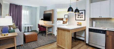 A modern hotel room with a wooden floor, a kitchenette, a dining area, and a living space with a sofa and a desk.