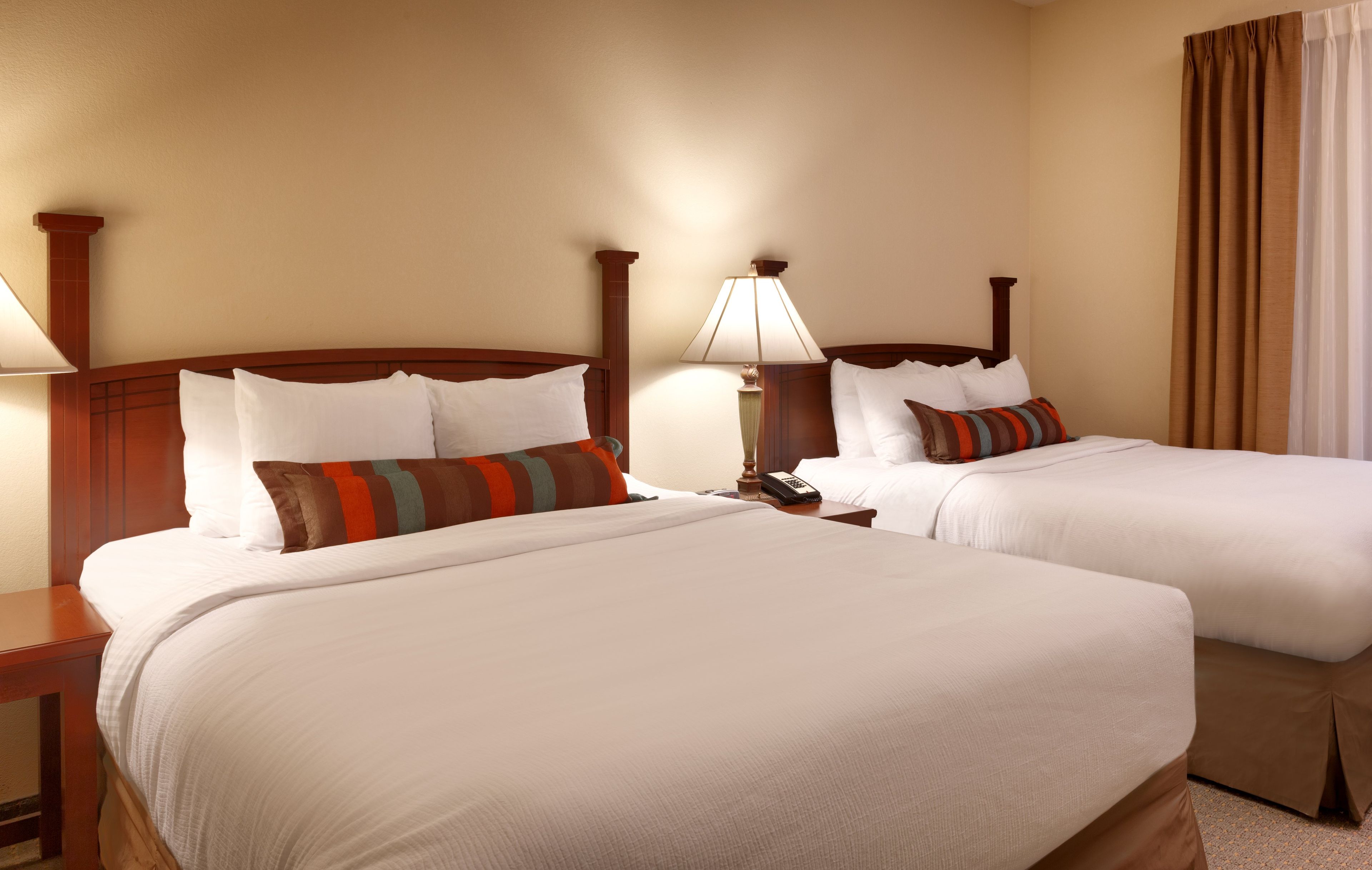 A hotel room with two beds, each with white linens and multiple pillows. There is a bedside table with a lamp and a telephone.