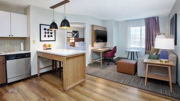 A modern hotel room with a kitchen area, a dining table, a sofa, and a TV.