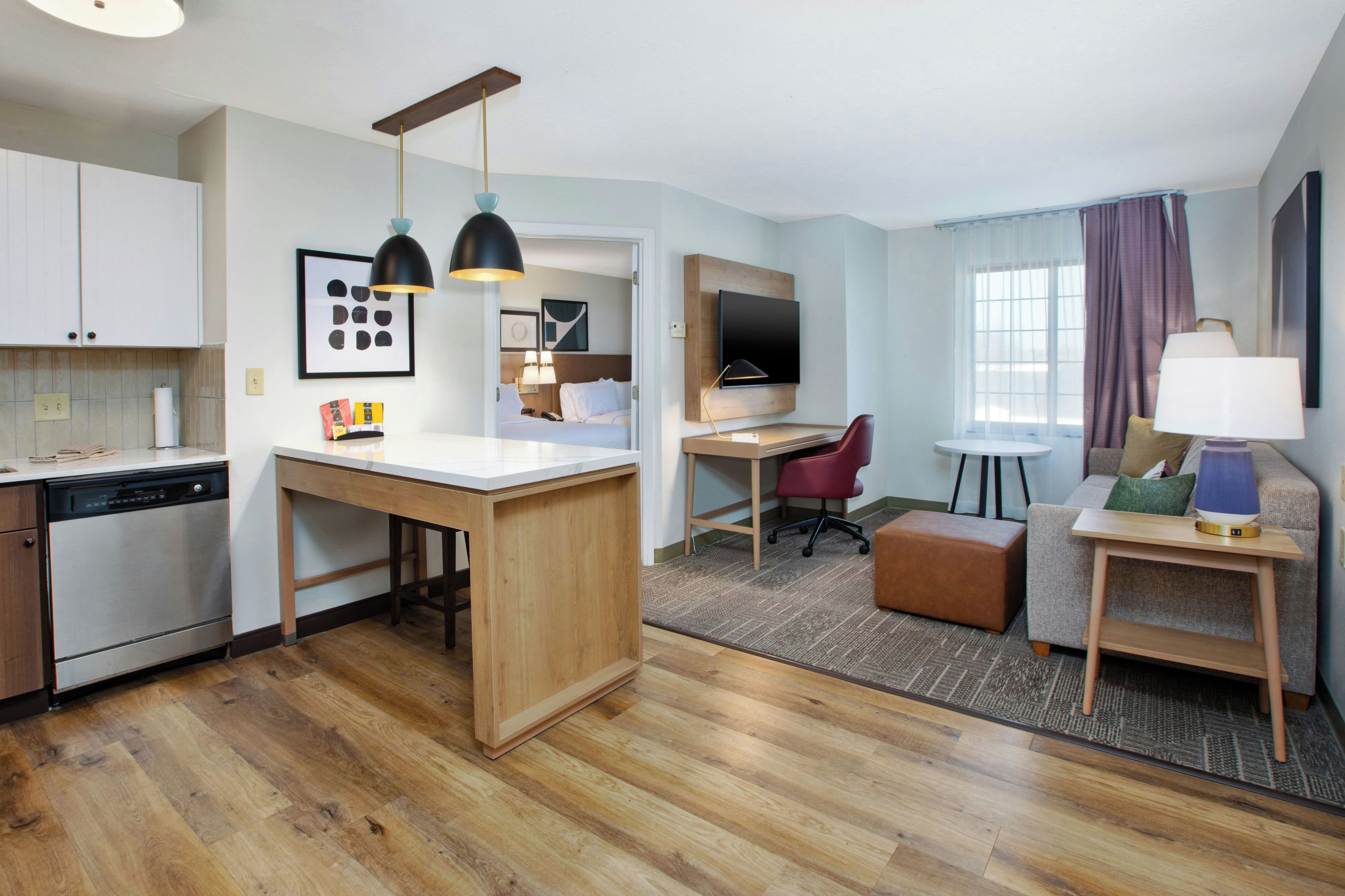 A hotel room with a kitchenette, a dining area, a workspace, and a seating area with a TV and lamp.