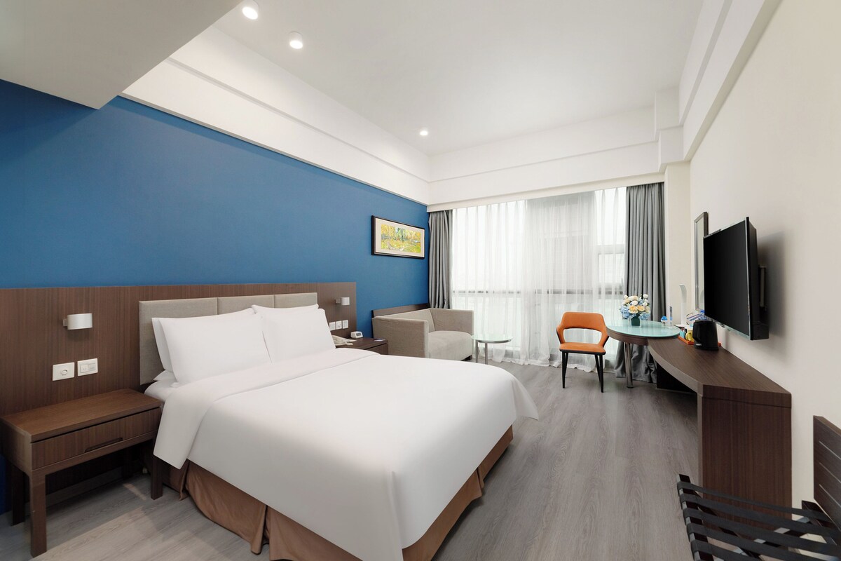 A hotel room with a large bed, a desk with a chair, a TV, and a window with curtains.