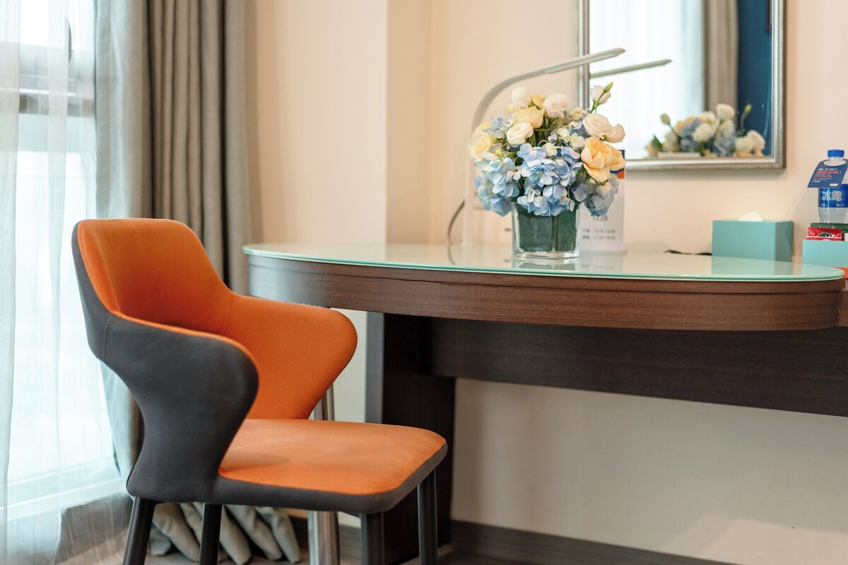 A modern hotel room with a glass-top desk, a chair, a vase of flowers, and a mirror.