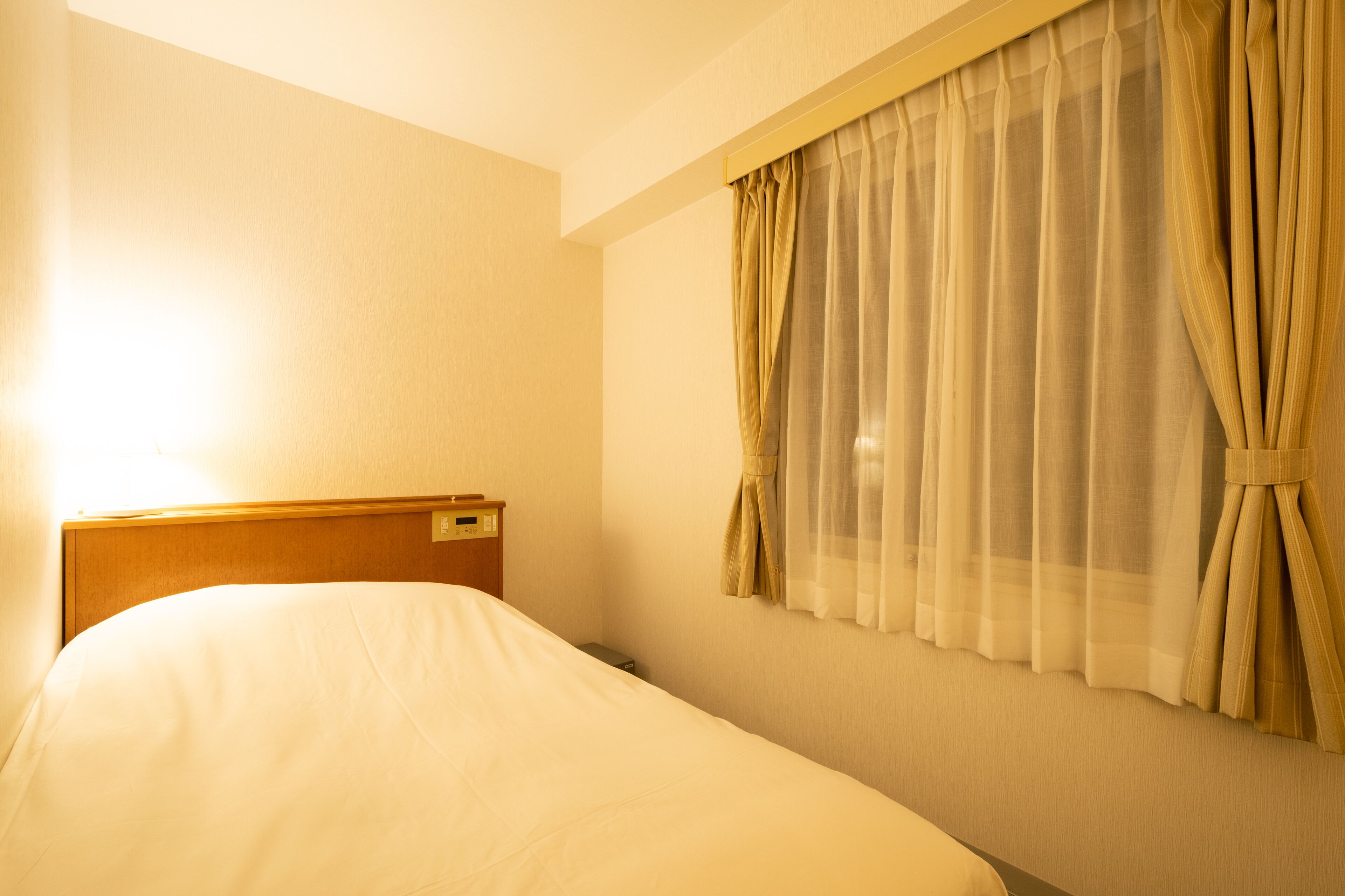 A hotel room with a single bed, a nightstand, and a window with sheer curtains.