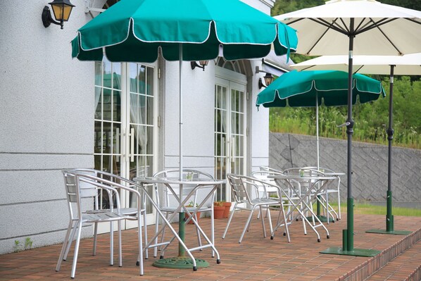 Des chaises et des tables pour terrasse, avec des parasols, un bâtiment blanc doté de fenêtres et un chemin pavé de briques.