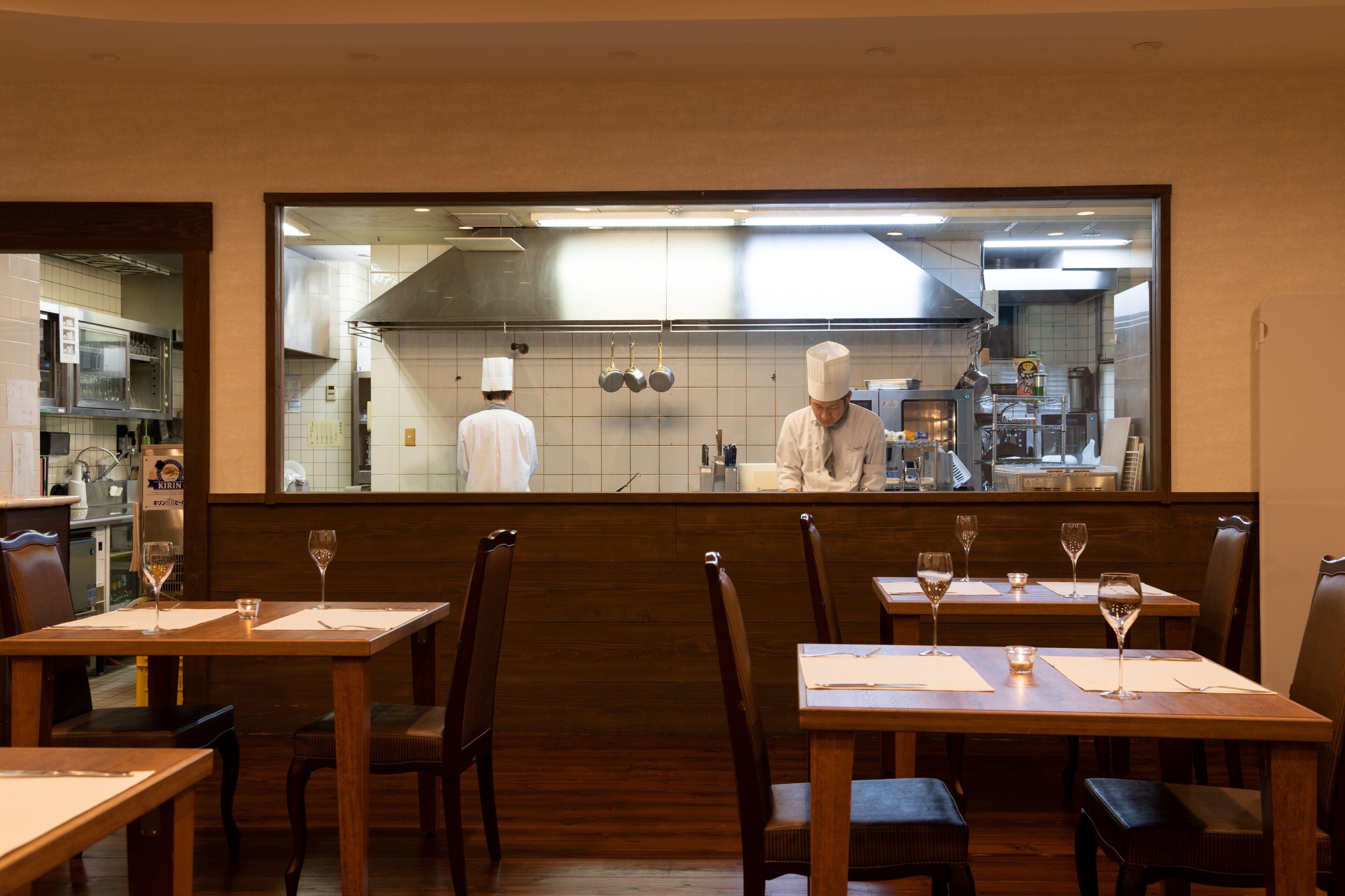 Un restaurante con mesas y sillas de madera, un chef en la cocina y vistas a la cocina a través de un separador de vidrio.