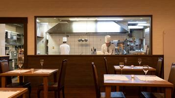 Un restaurante con mesas y sillas de madera, un chef en la cocina y vistas a la cocina a través de un separador de vidrio.