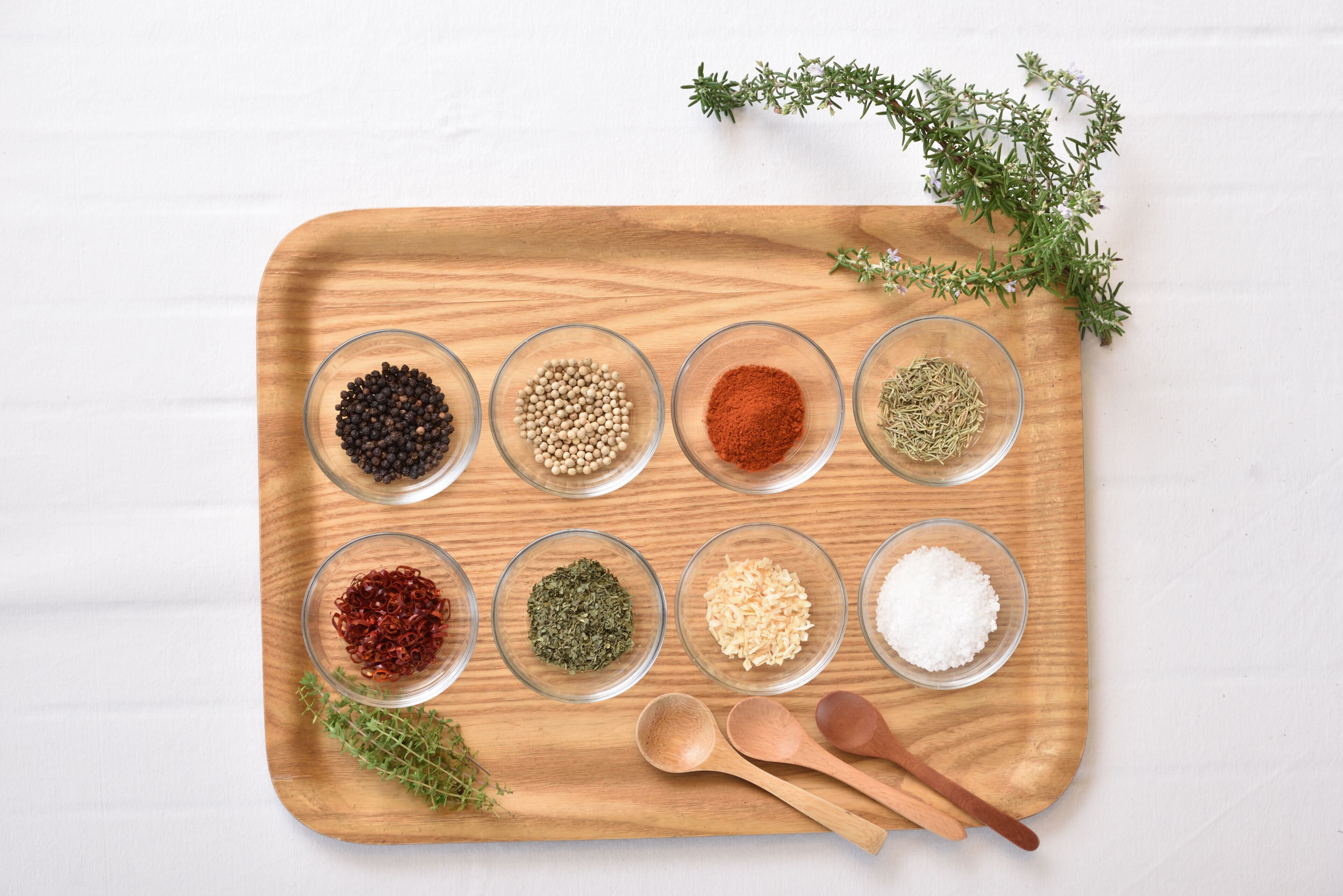 A wooden tray with glass bowls containing various spices and herbs, accompanied by wooden spoons and fresh thyme.