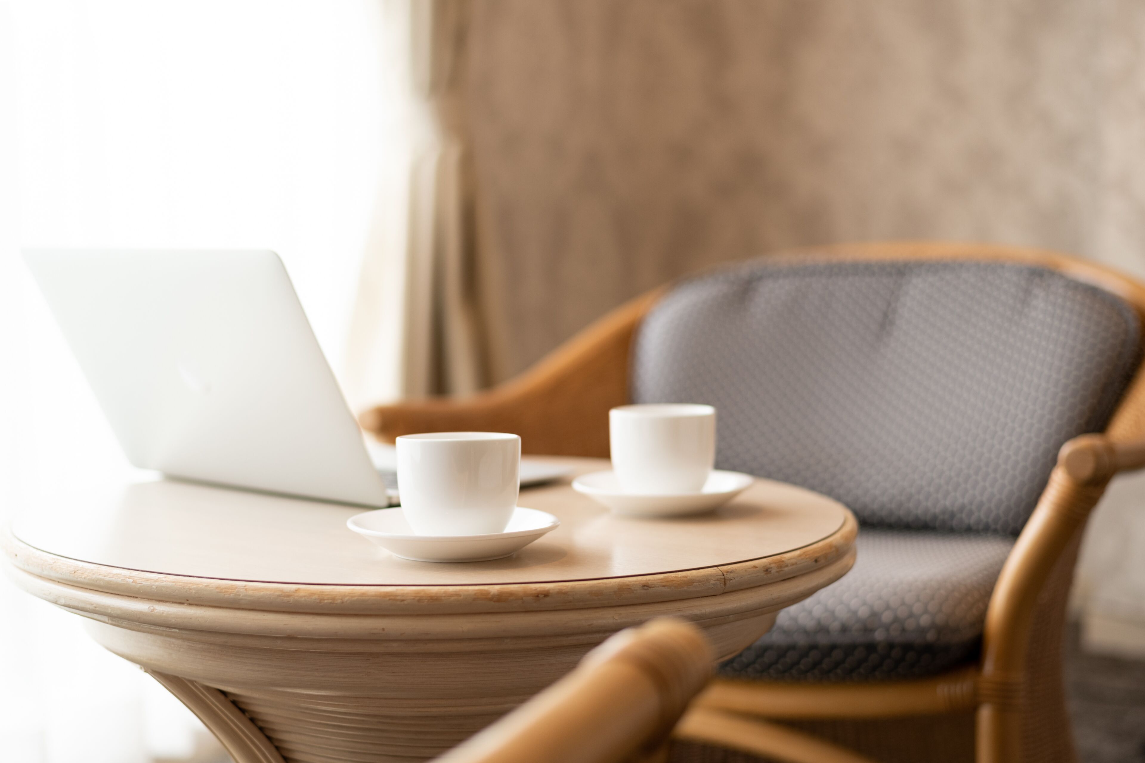 A wicker chair with a laptop and two white cups on a round table.