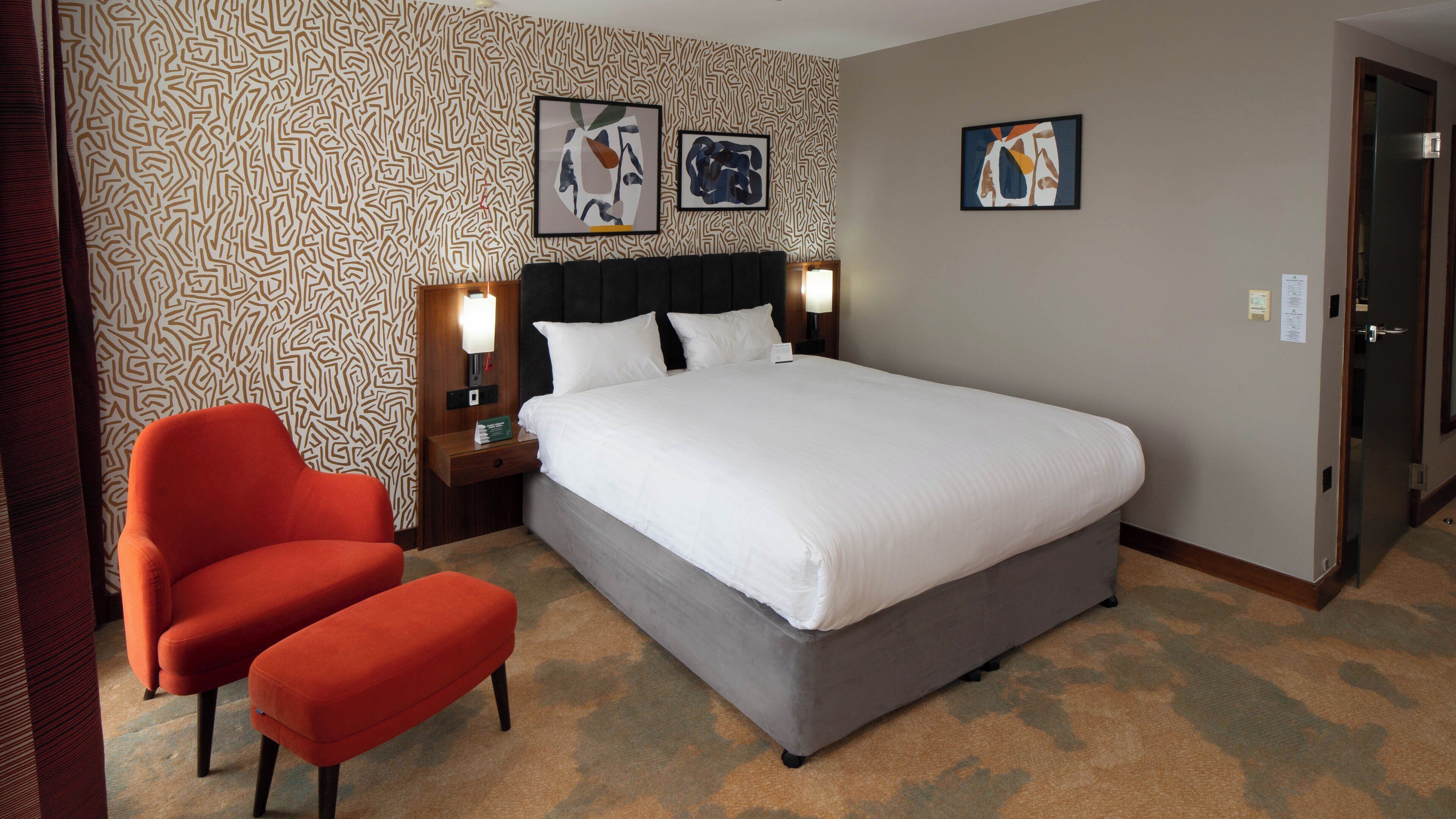 A hotel room with a large bed, two bedside tables with lamps, a red armchair, and patterned wallpaper.