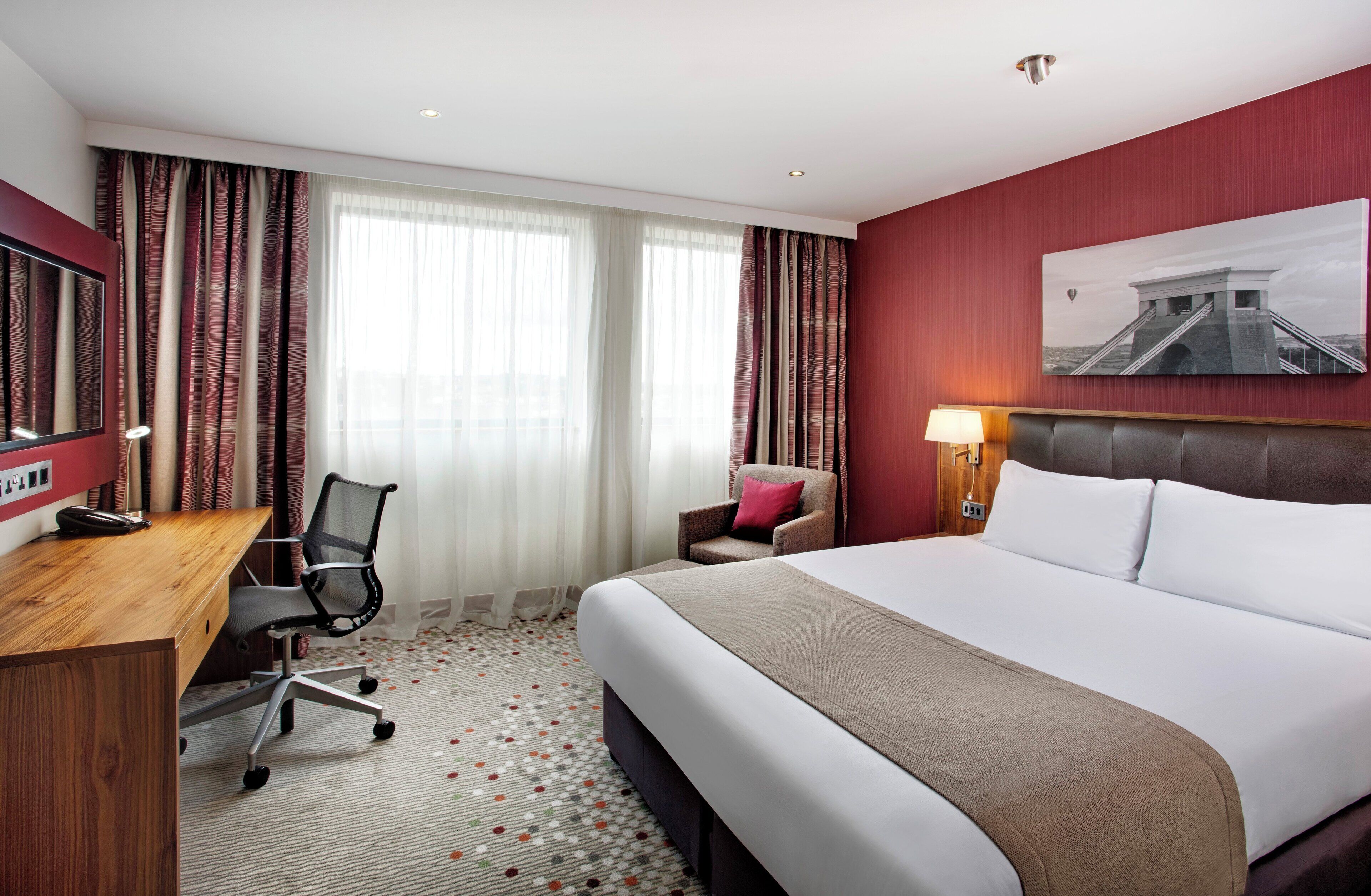 Una habitación de hotel con cama, escritorio, silla y un cuadro en la pared.