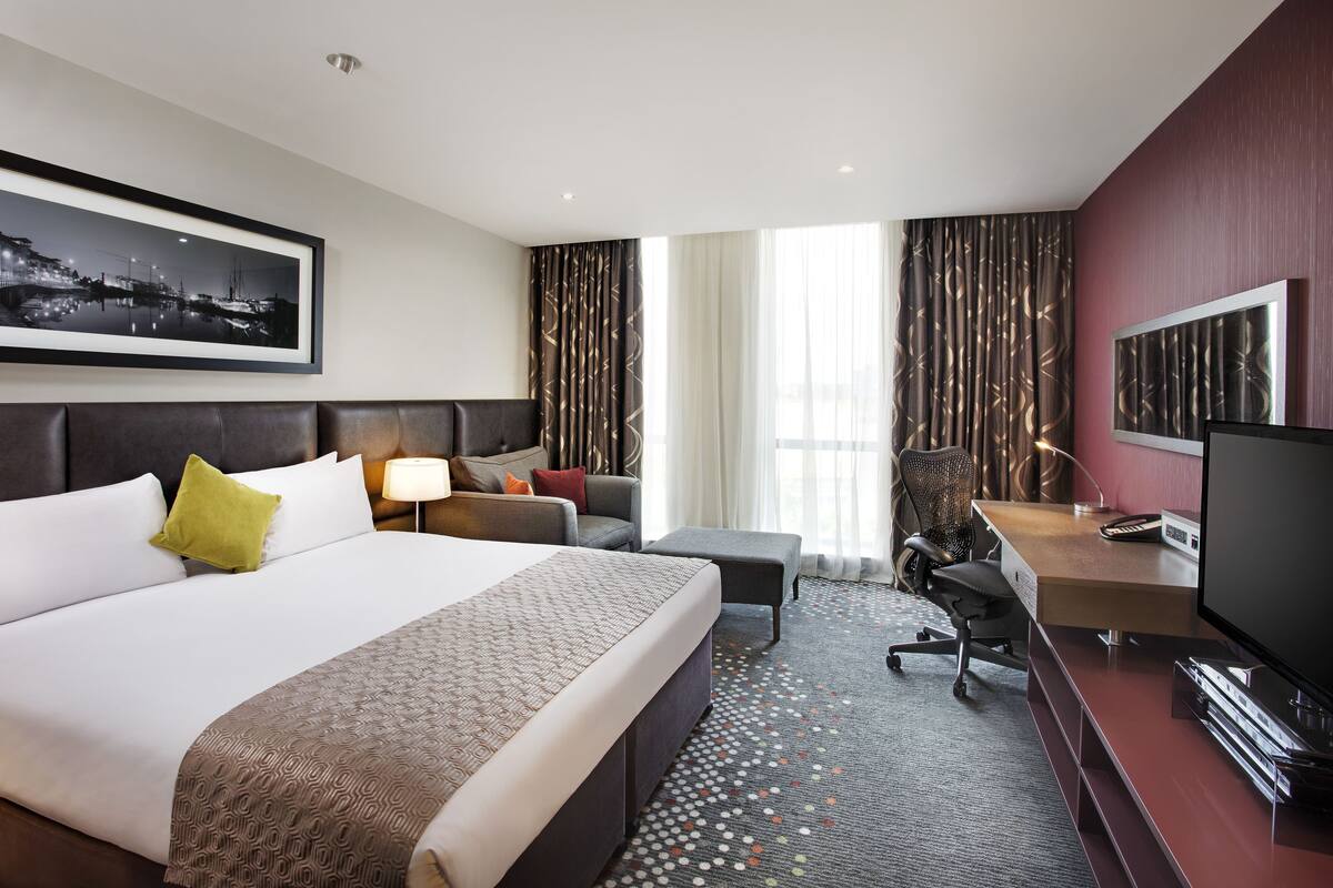A hotel room with a large bed, a desk with a TV, a chair, and a sofa.