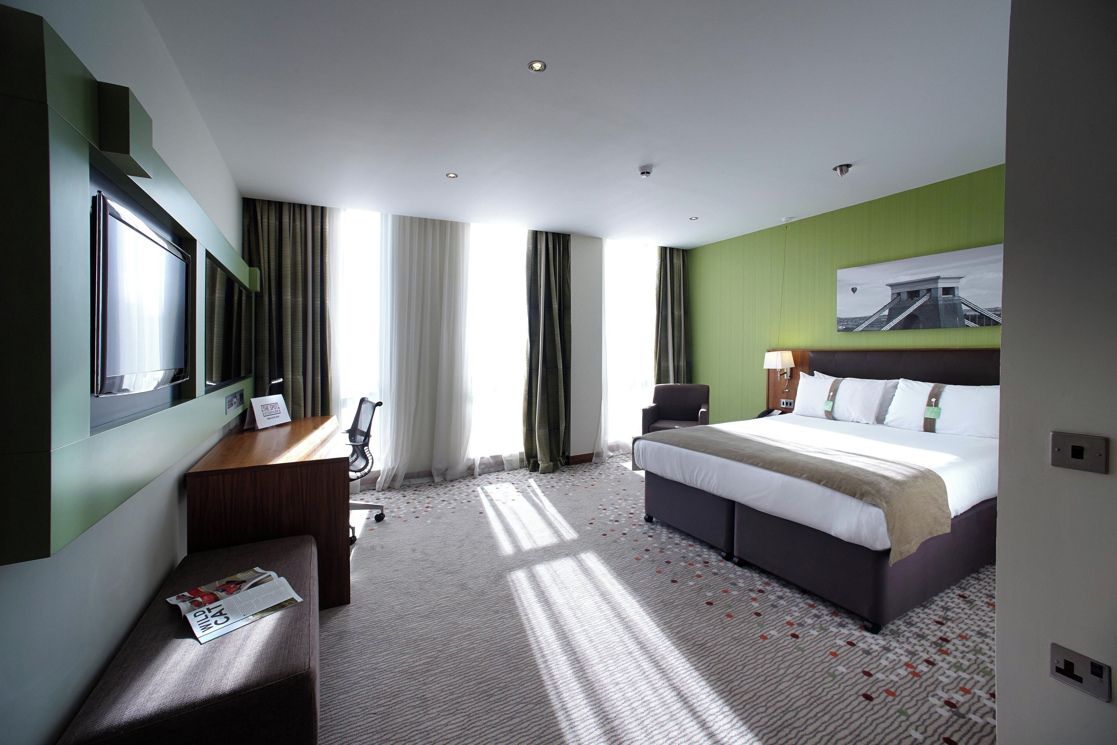 A hotel room with a bed, a desk, a chair, a window with curtains, and a painting on the wall.