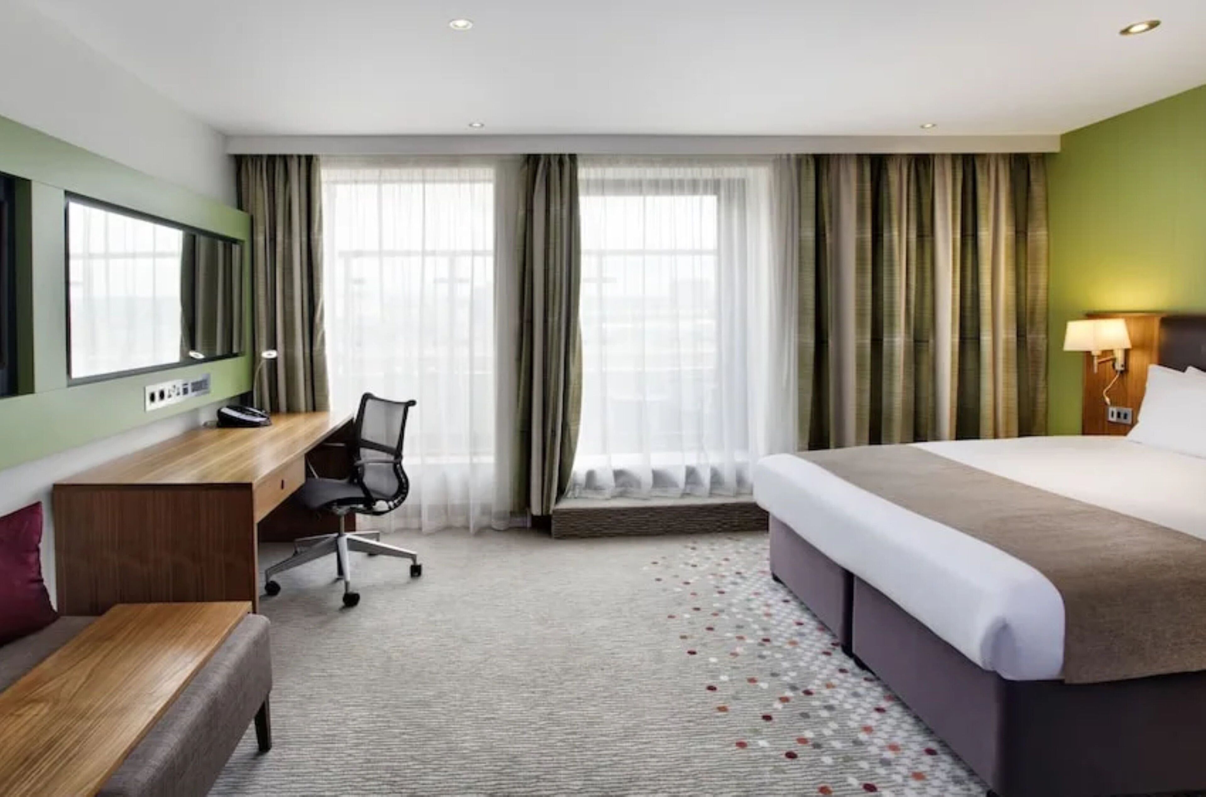 Een hotelkamer met een bed, bureau, stoel, groot raam met gordijnen en een tv aan de muur.