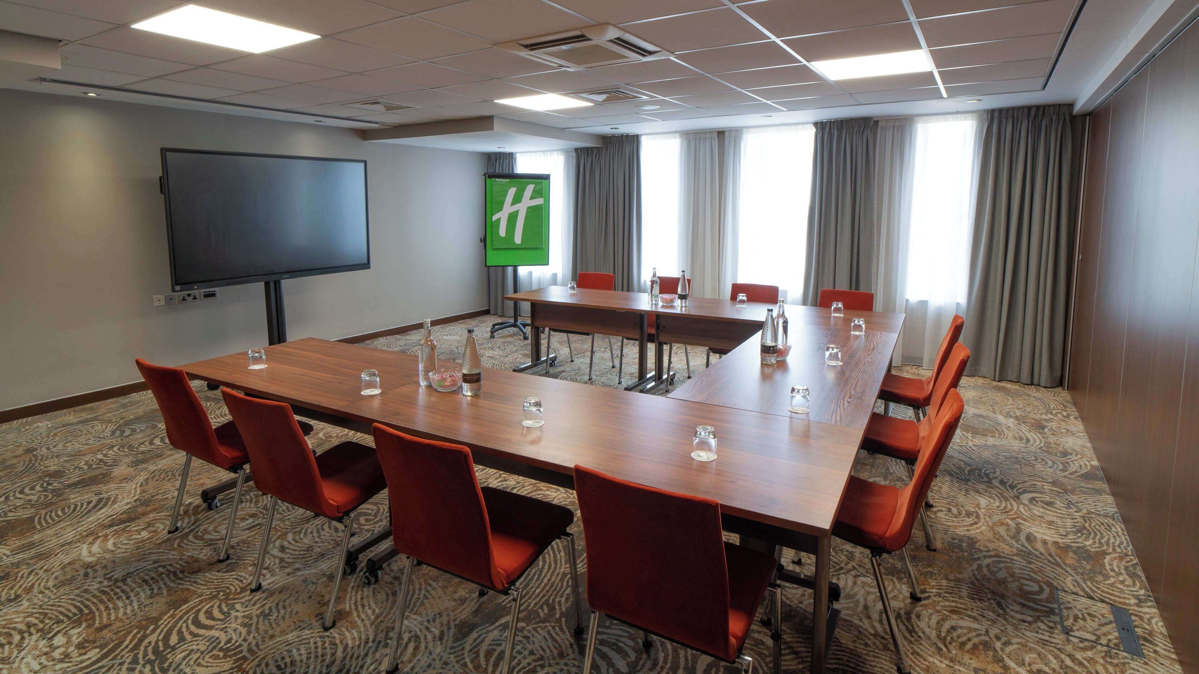 Een vergaderruimte met een grote tafel, rode stoelen, een projectiescherm en een groen bord aan de muur.