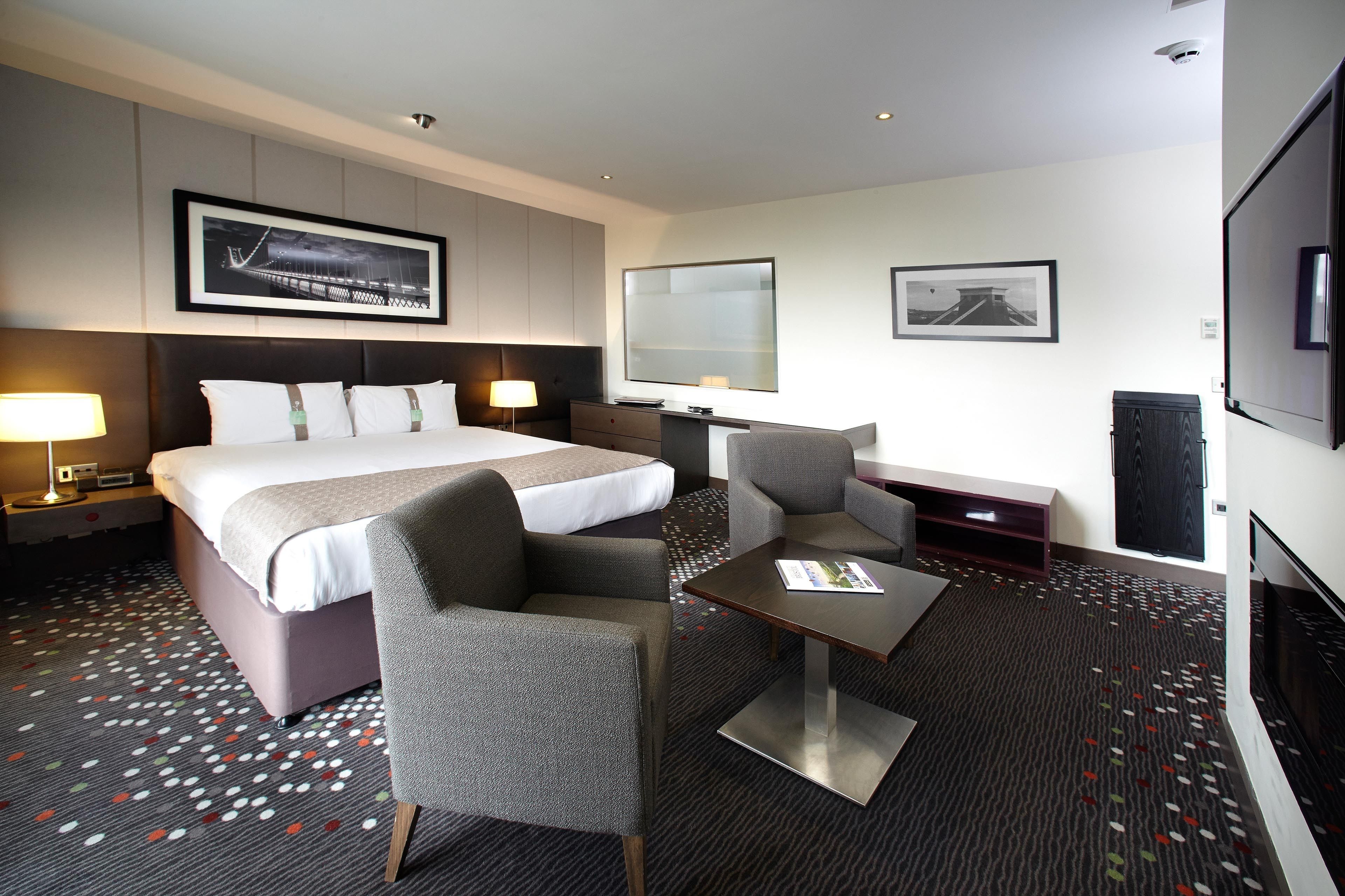 A hotel room with a bed, two armchairs, a coffee table, a TV, and a large window.