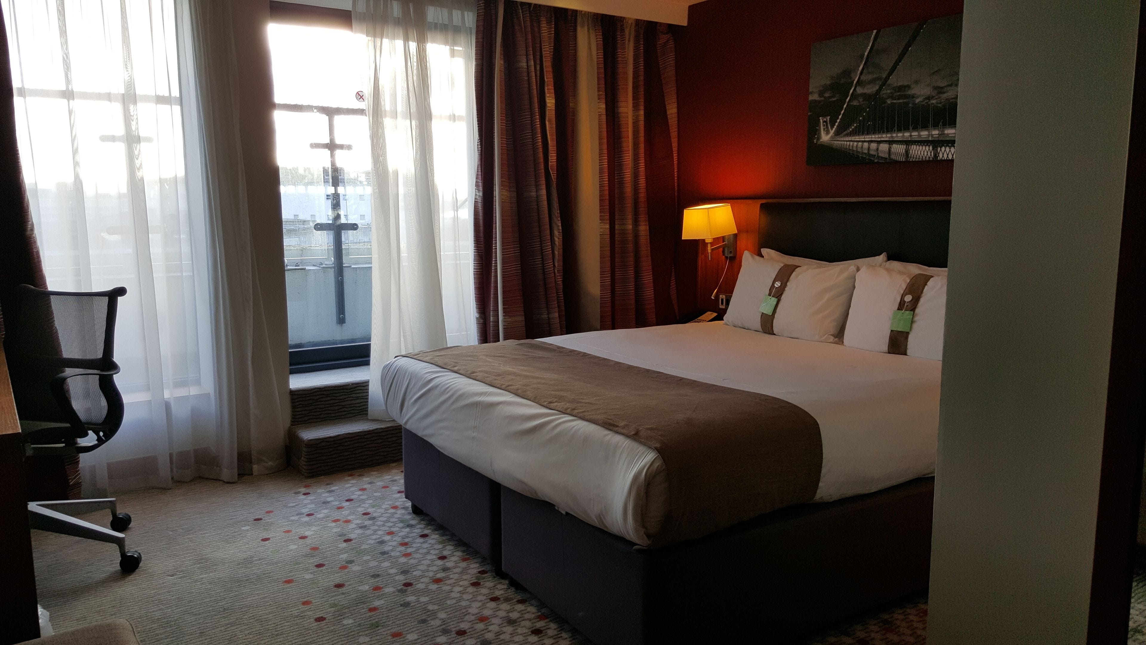 A hotel room with a bed, a desk, and a chair. There is a window with curtains, a lamp, and a painting on the wall.