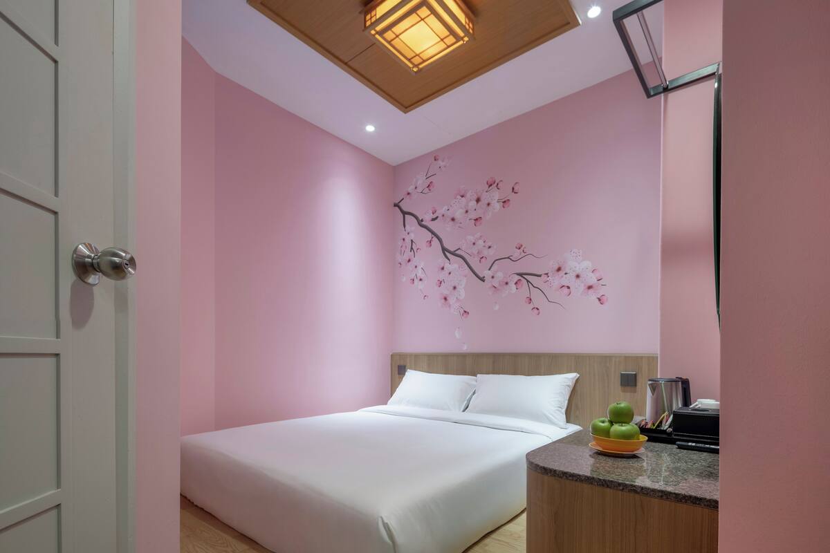 Quarto com cama, cabeceira de madeira, parede com mural de cerejeiras e pequena área de cozinha com uma tigela de frutas.