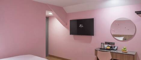 Una habitación de hotel con paredes rosas, un televisor montado en la pared, un escritorio con una silla y dos camas.
