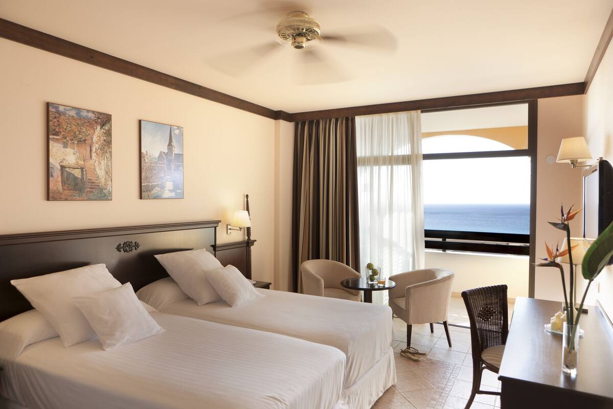 A hotel room with two beds, a dining area, and a balcony with a view of the sea.