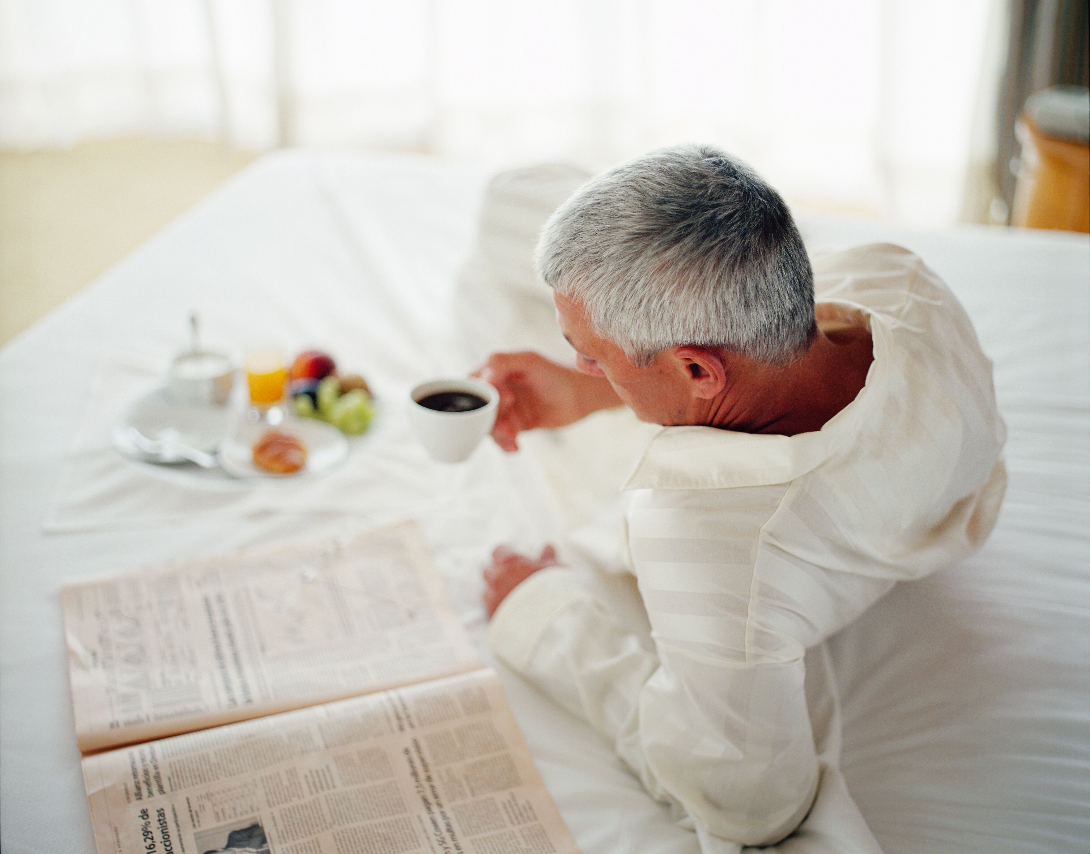 A person lying on a bed with a newspaper, a cup of coffee, and a plate of fruit.