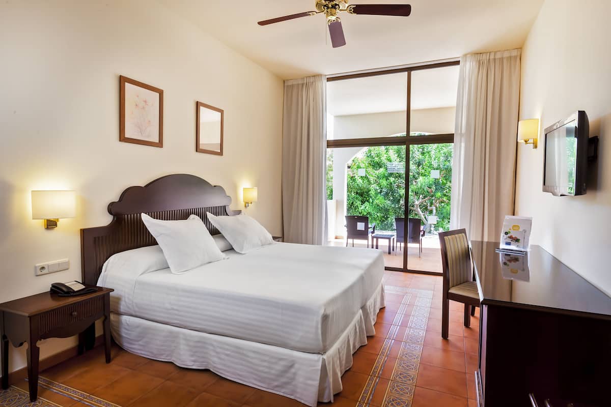 A hotel room with a large bed, bedside lamps, a desk with a television, and a view of greenery outside.