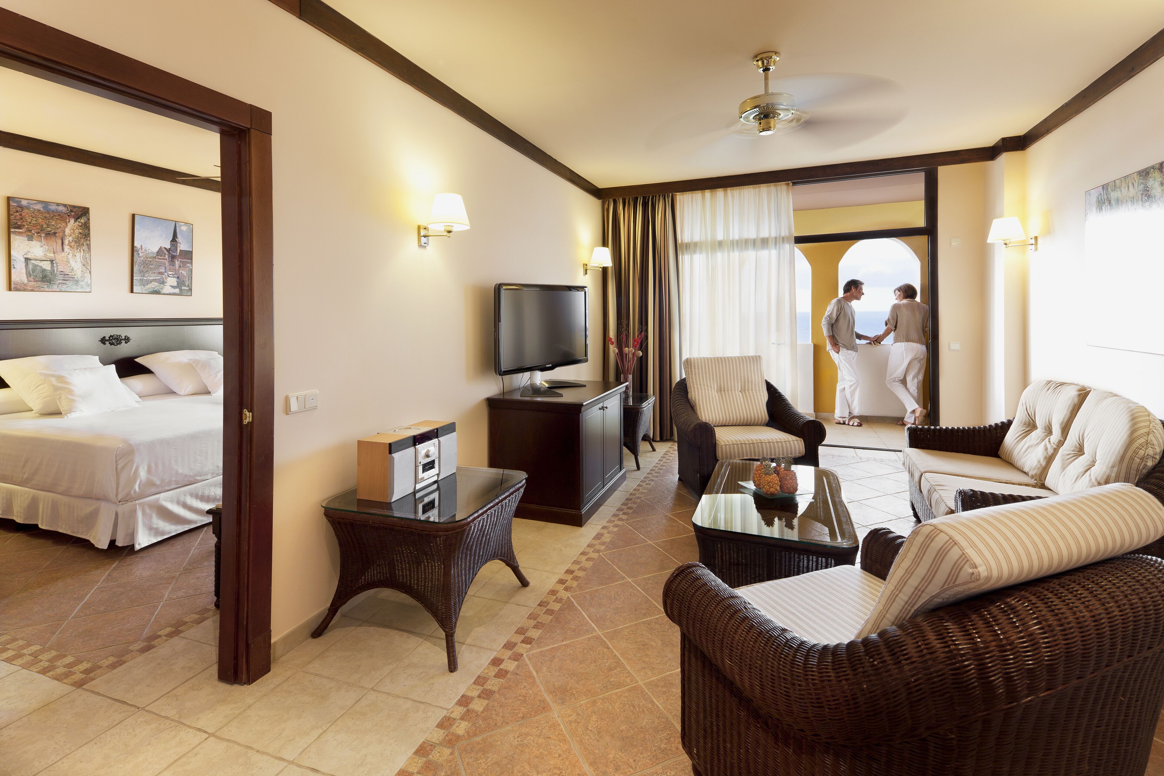 A hotel room with a bed, a TV, wicker furniture, and a view of a couple outside.