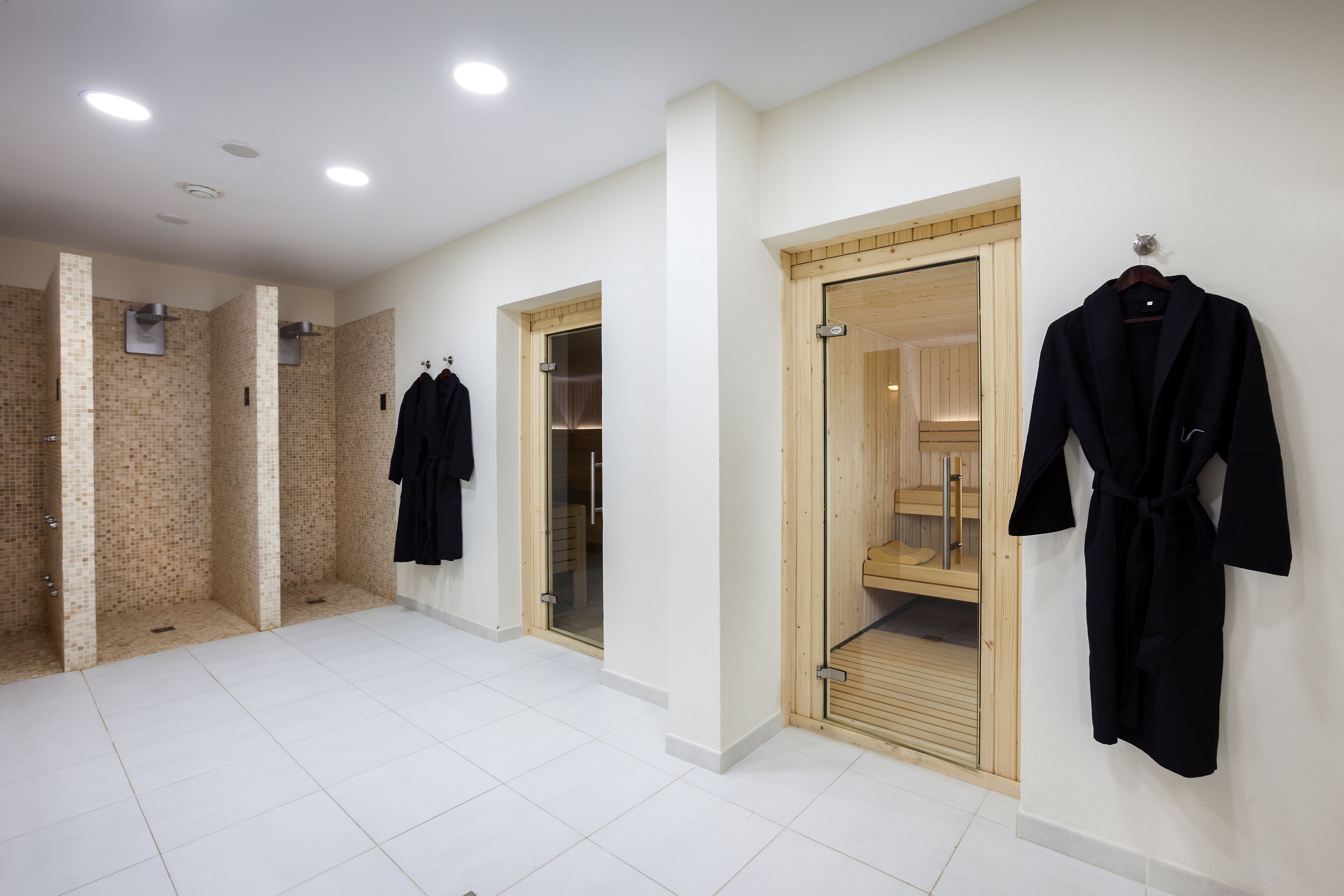 A spacious bathroom with white tiled floors, a wooden sauna, and black robes hanging on the wall.