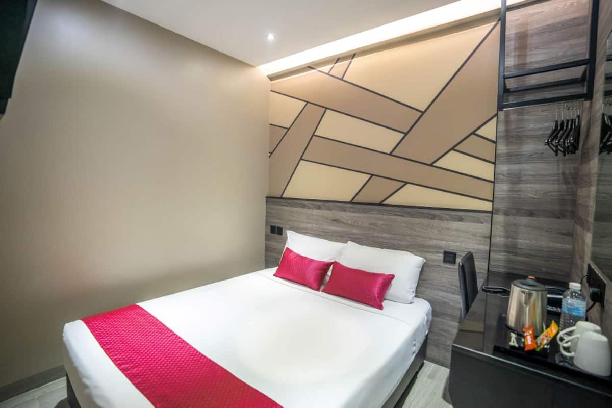 Quarto de hotel moderno com uma cama grande, cabeceira com padrões geométricos, mesa de cabeceira com chaleira e um gancho na parede para chaves.