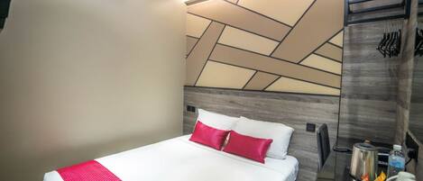 A modern hotel room with a large bed, a headboard with geometric patterns, a bedside table with a kettle, and a wall-mounted hook for keys.