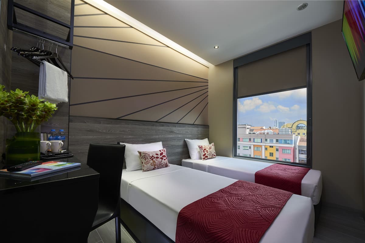 Quarto de hotel com duas camas, uma escrivaninha com um laptop, um vaso com plantas e vista para edifícios coloridos.