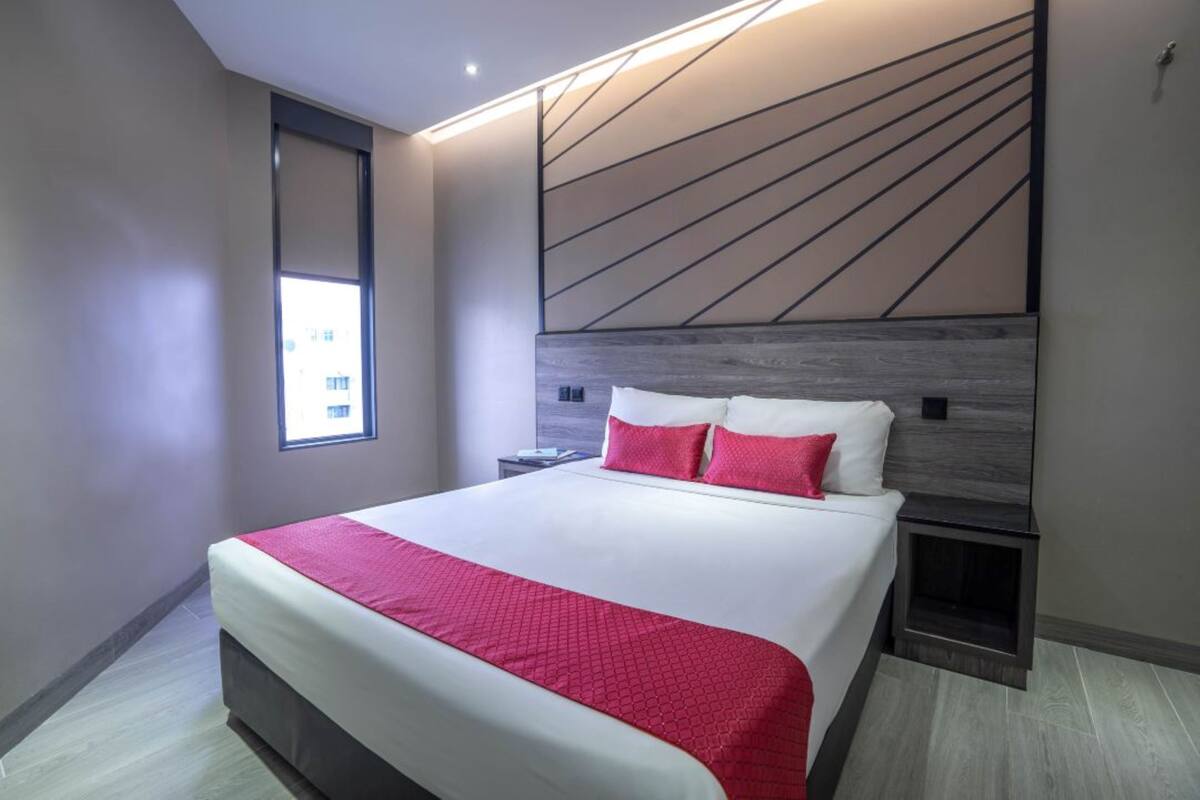 Quarto de hotel moderno com uma cama grande, uma mesa de cabeceira, televisão integrada e uma janela com vista para edifícios.