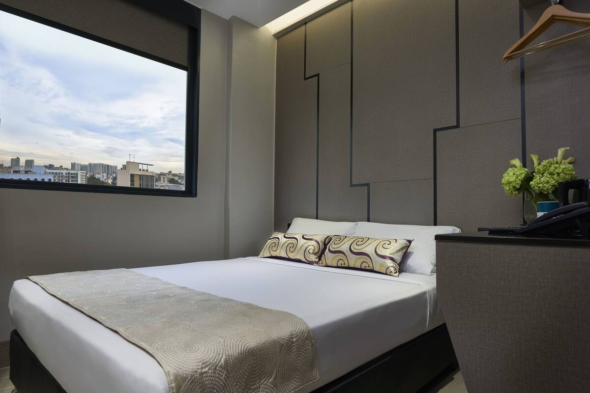 Quarto de hotel moderno com uma grande janela, cama com almofadas estampadas, mesa de cabeceira e vista para a cidade.