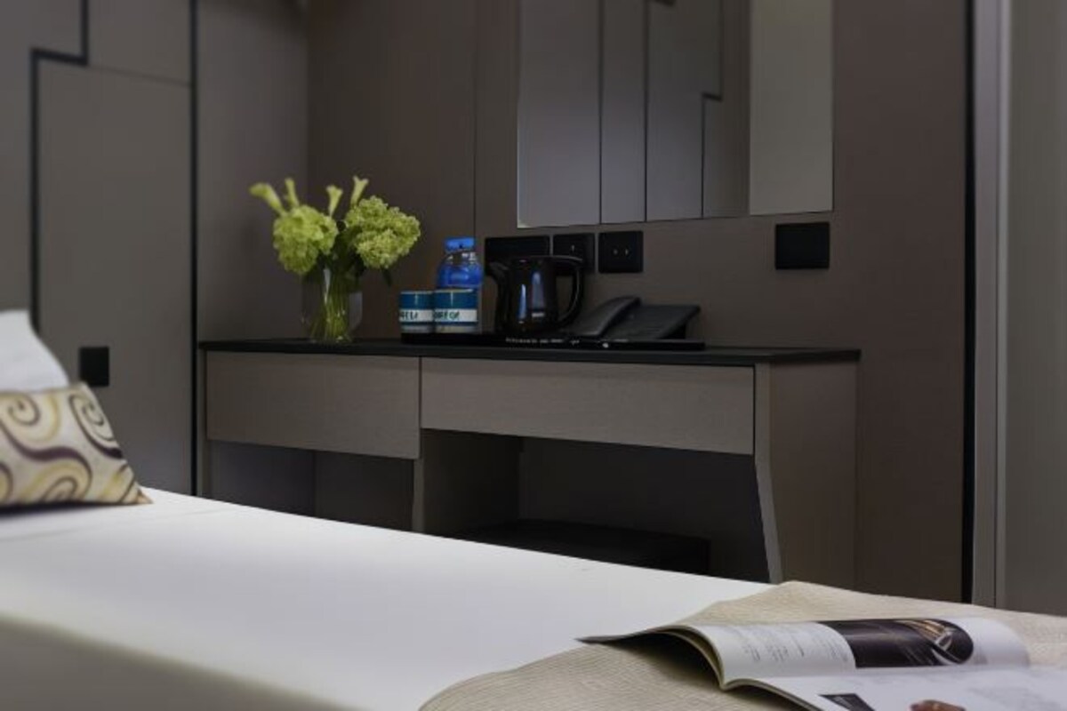 A hotel room with a bed, a bedside table with a vase of flowers, a water bottle, a coffee maker, and a telephone.