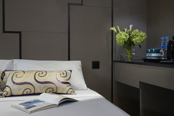 Una cama bien hecha con un cojín estampado, un libro y un florero con flores sobre una mesita de noche.