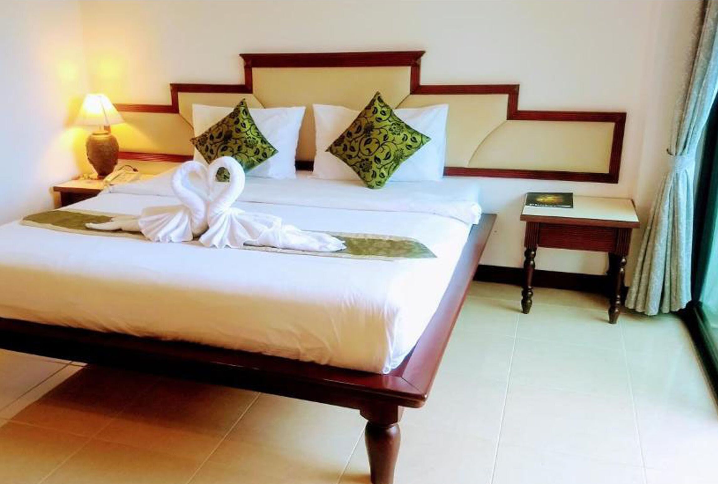 A neatly made bed with a white duvet, two green patterned pillows, and a white towel arranged in a swan shape on the bed.