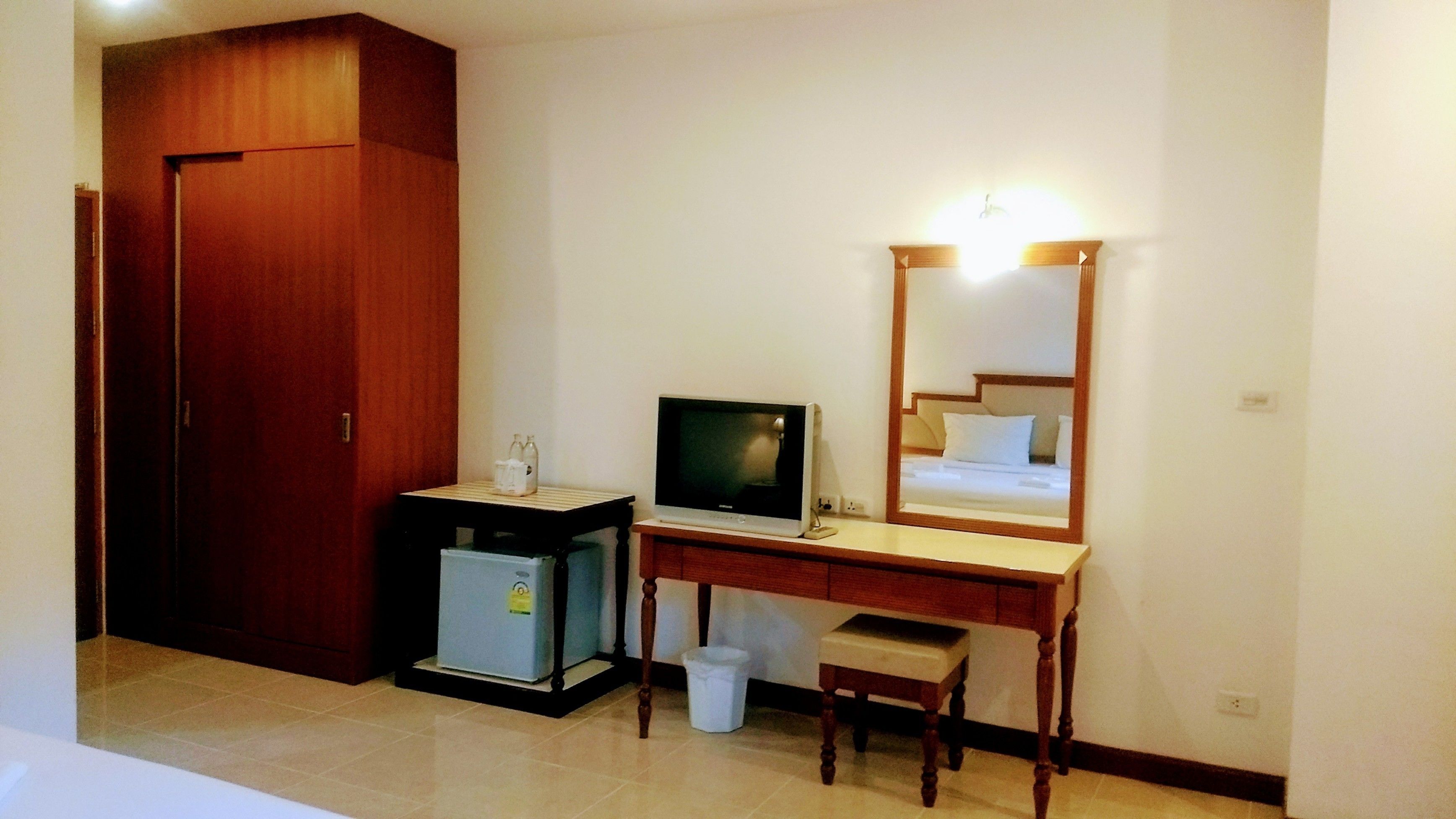 A hotel room with a wooden wardrobe, a desk with a television, a small trash bin, and a mirror.