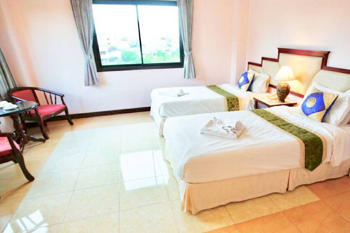 A hotel room with two beds, a small table with a chair, a window with curtains, and a lamp on the nightstand.