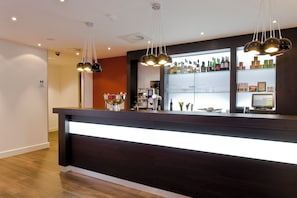A bar area with a dark counter, overhead lighting, and shelves with bottles and glasses. - Holiday Inn Express Amsterdam - Schiphol by IHG (Hoofddorp)